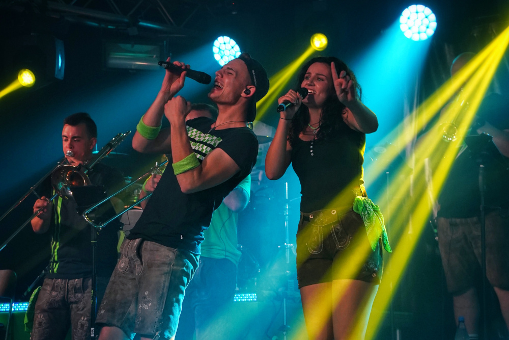 Sänger und Sängerin der Partyband mit Posaunen im Hintergrund und gelbem Scheinwerfer-Licht